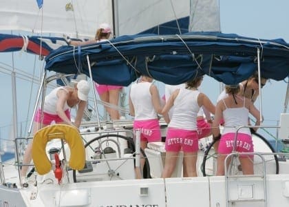Multiple guests gathered on the deck of a catamaran under a blue canopy, anchored in clear waters with other yachts visible in the background