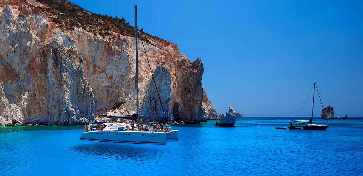 Catamaran anchored in turquoise water below towering limestone cliffs, with guests on deck and other boats moored nearby