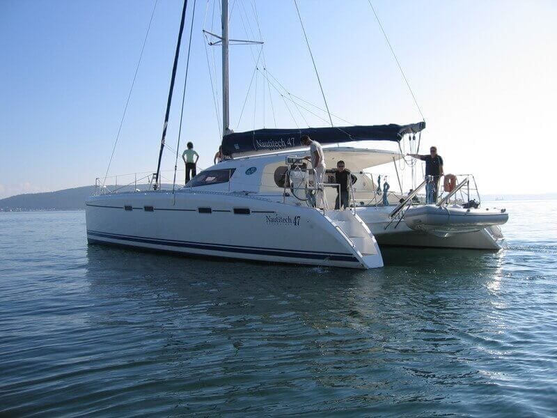 White catamaran at anchor with guests and crew visible on deck and tender alongside in calm blue water