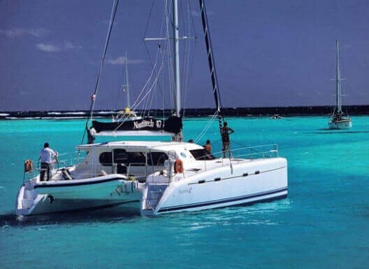 White catamaran anchored in turquoise water with guests on deck, other sailboats visible in background