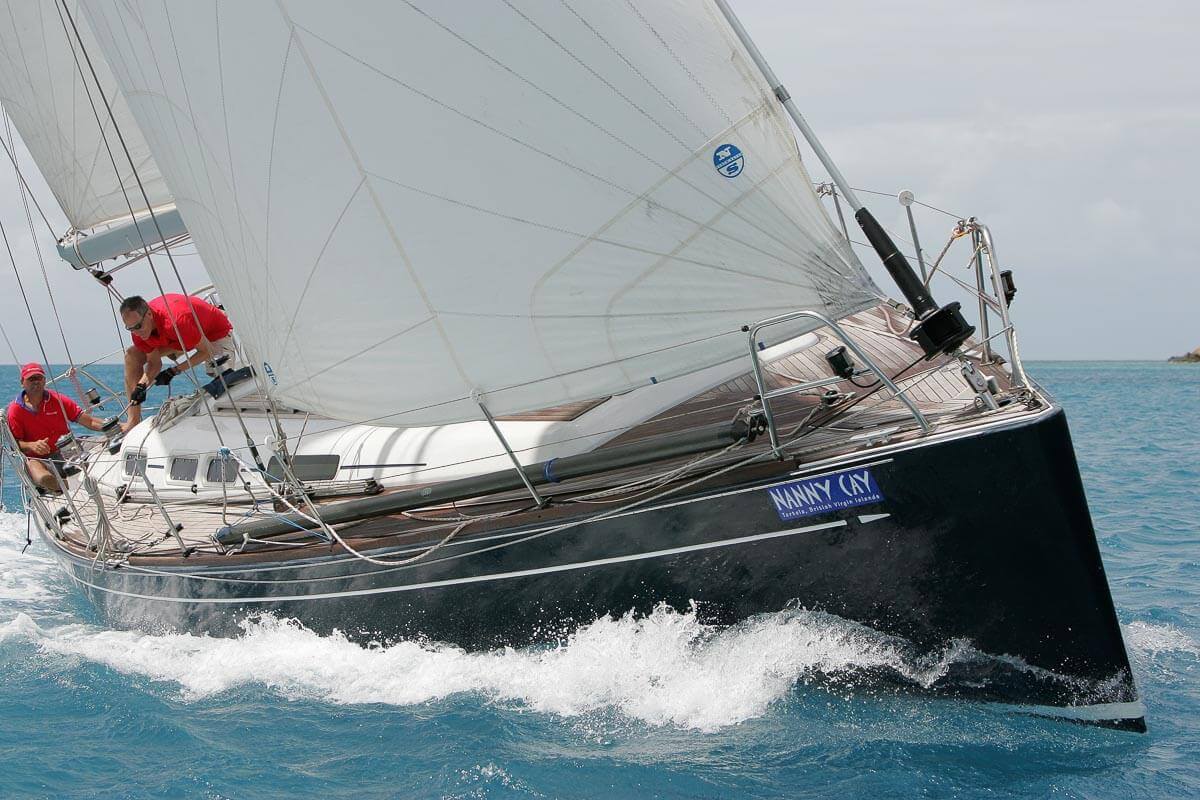 Monohull yacht with mainsail raised, crew in red shirts managing sails near shore in shallow turquoise water