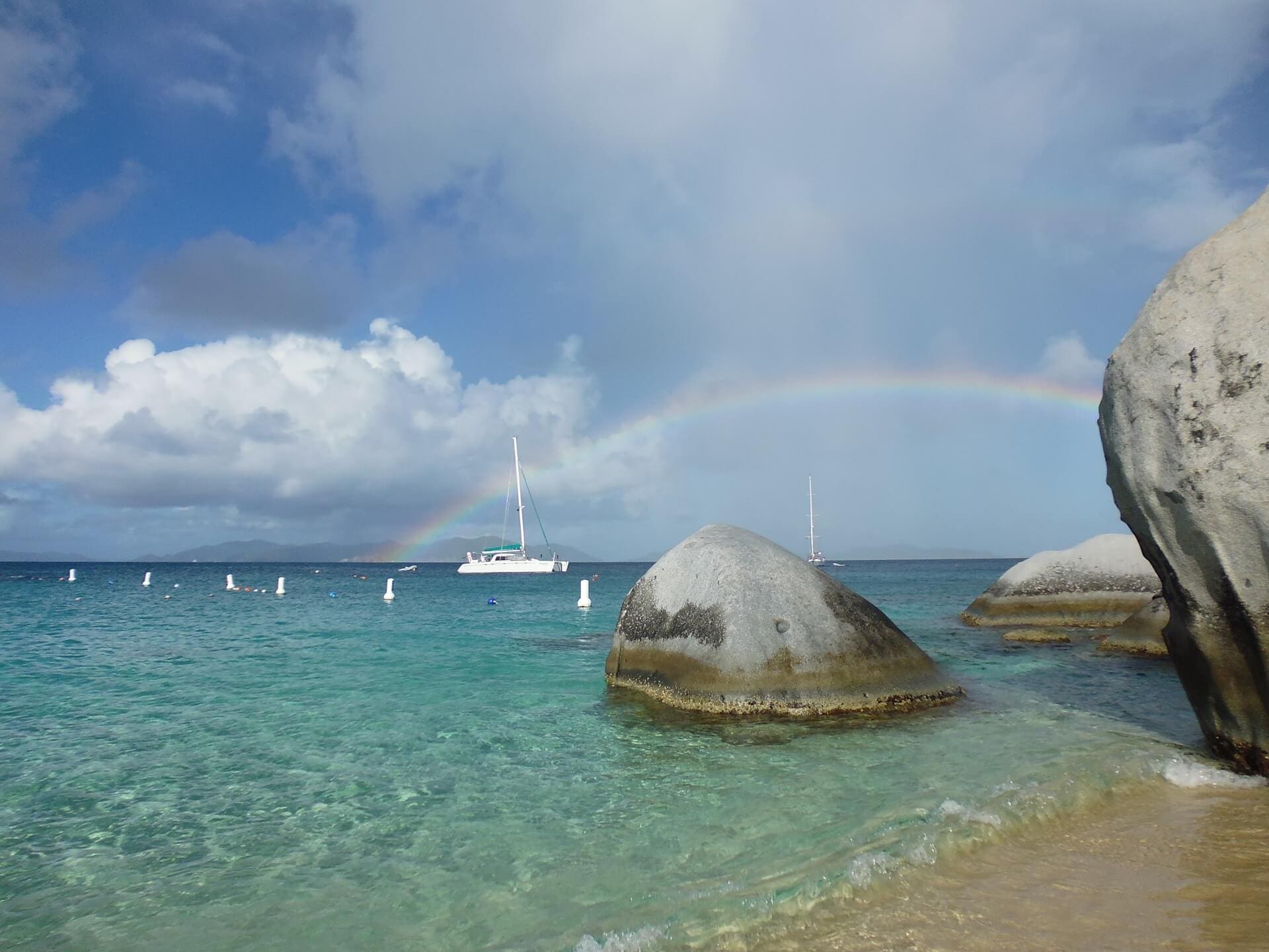 Catamaran anchored in turquoise water near granite boulders, with rainbow visible in sky above distant island