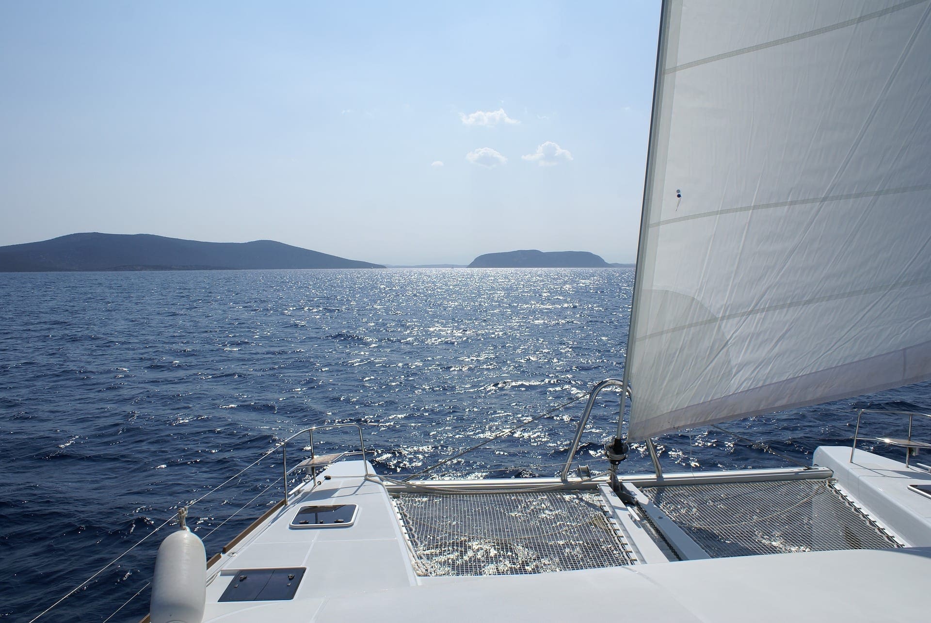 Catamaran with mainsail raised sailing past rocky islands in clear blue water under light clouds